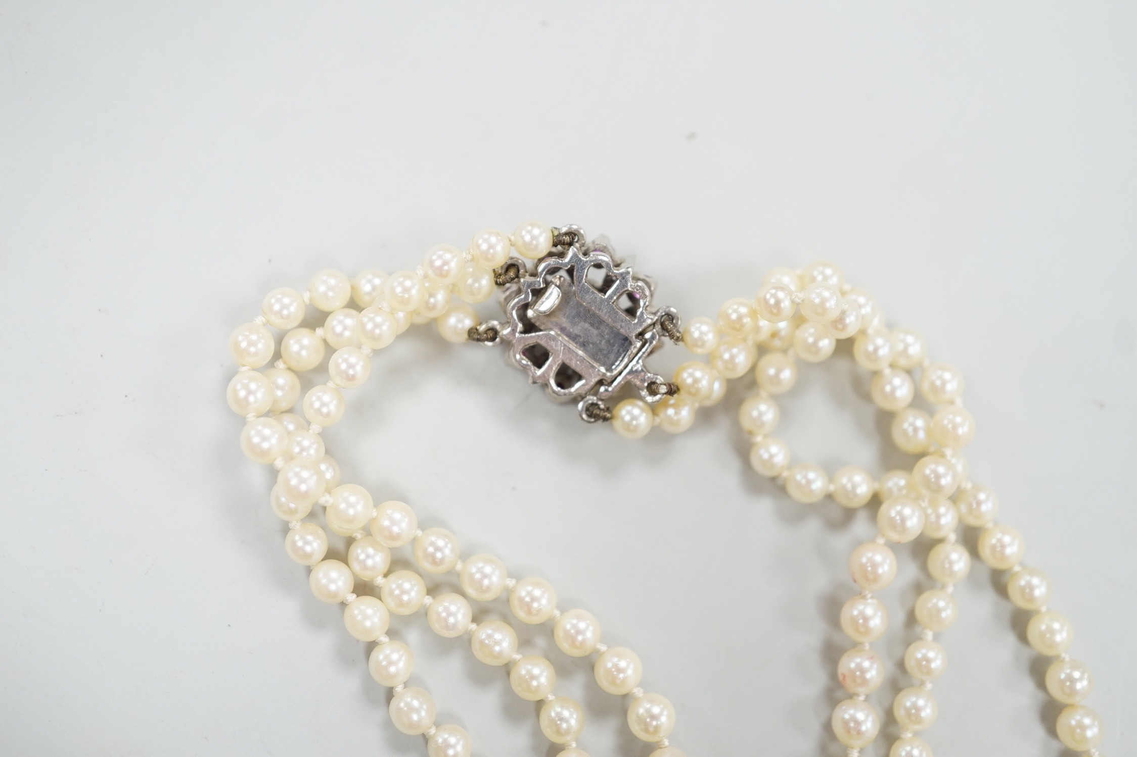 A 1960's triple strand graduated cultured pearl necklace, with 9ct white gold, ruby and diamond chip cluster set clasp, 49cm.